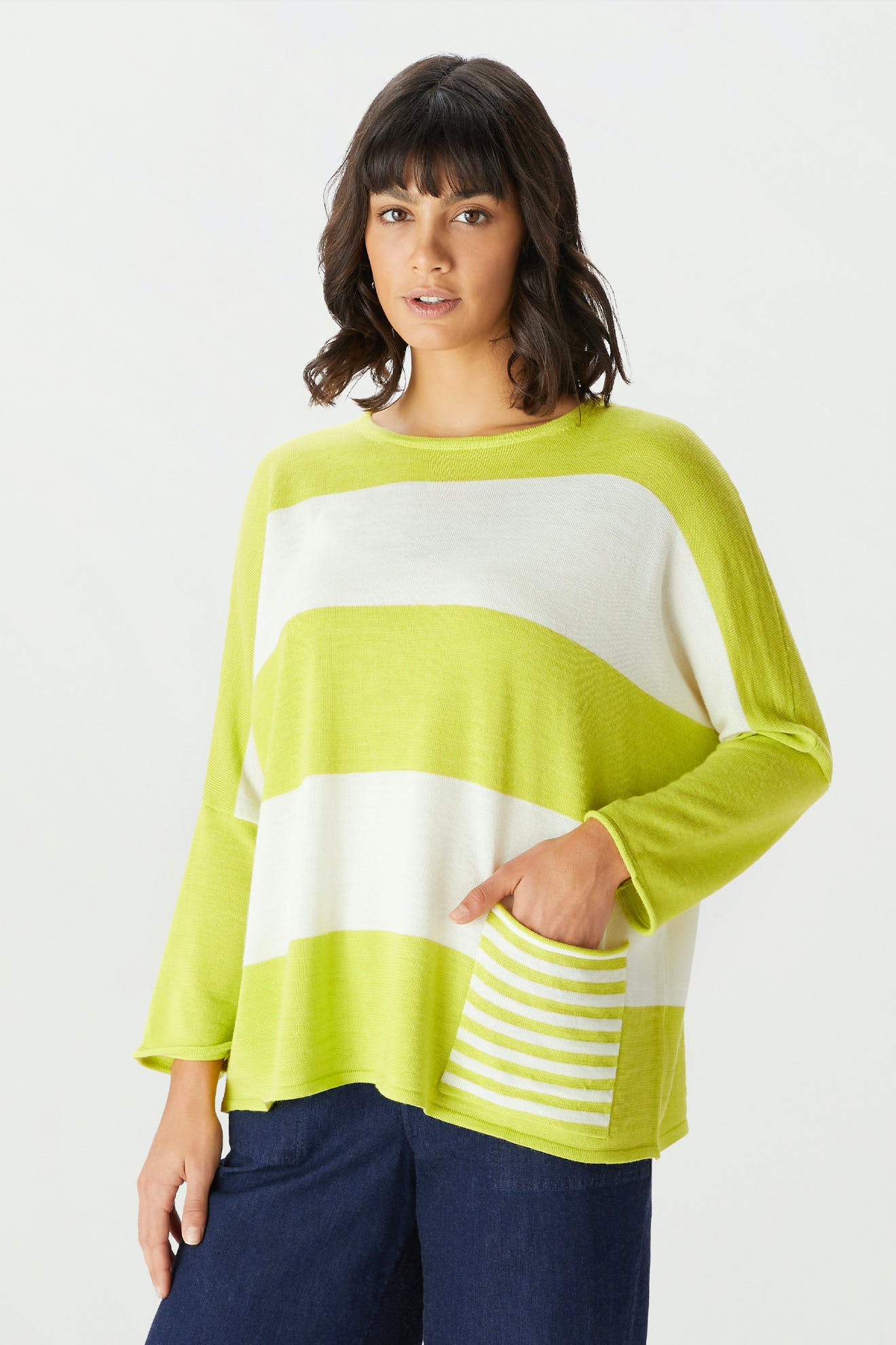A woman is wearing the Merino Blend Stripe Sweater, a loose-fitting lime green and white striped knit featuring a striped pocket, paired with dark blue pants and set against a plain light background—making it an ideal transitional weather piece for effortless style and comfort.