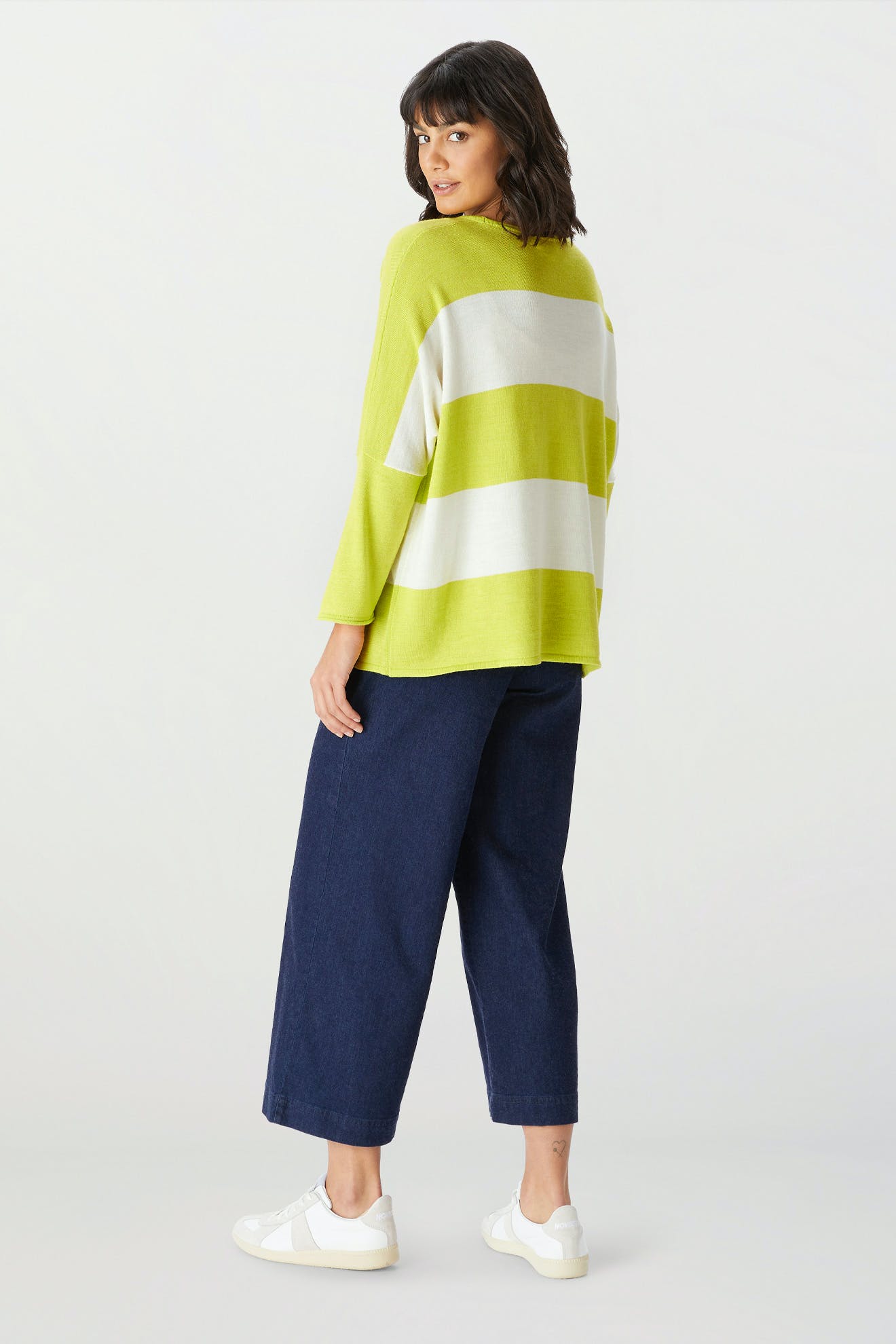 A woman stands facing away, wearing the Merino Blend Stripe Sweater—an ideal choice for transitional weather—paired with wide-leg dark blue jeans and white sneakers against a plain background.