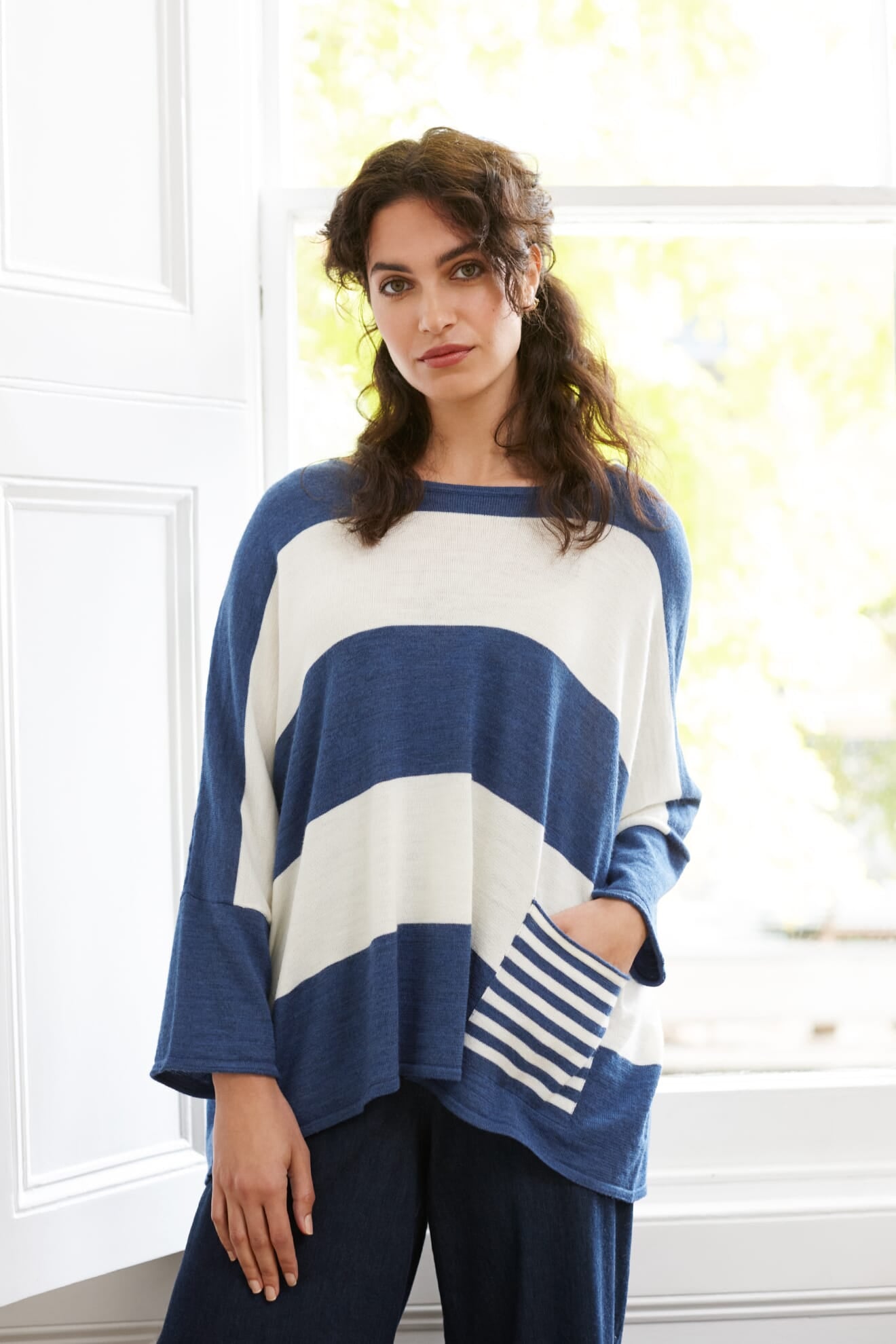 A woman stands indoors wearing the Merino Blend Stripe Sweater, featuring wide sleeves, a pocket, and loose blue and white stripes, paired with dark pants—a perfect transitional weather piece.