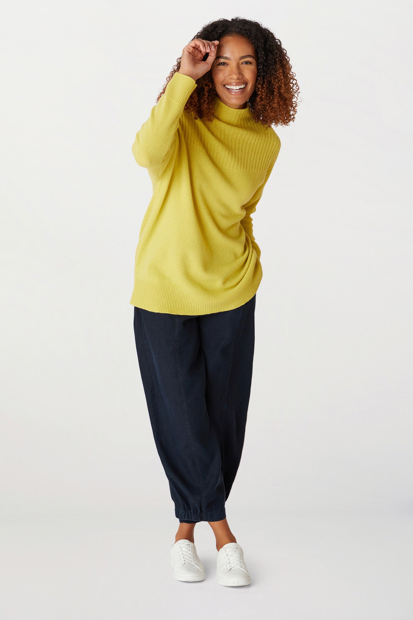 A woman with curly hair wearing a yellow Cashmere Blend Rib Turtle Neck, a classic cold-weather staple, navy blue pants, and white sneakers stands against a plain background, smiling and touching the brim of her sweater collar.