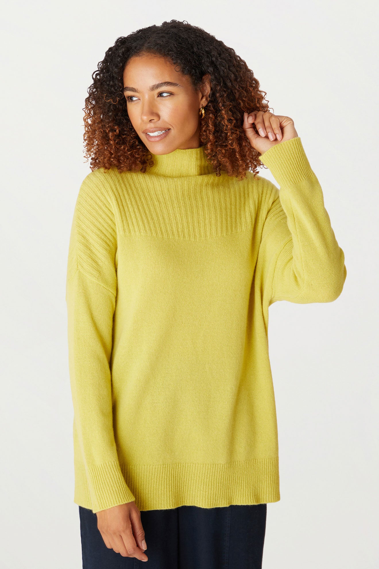 A woman with curly hair wears a bright yellow Cashmere Blend Rib Turtle Neck, a cold-weather staple, and black pants. She is standing against a plain background and looking to her right.
