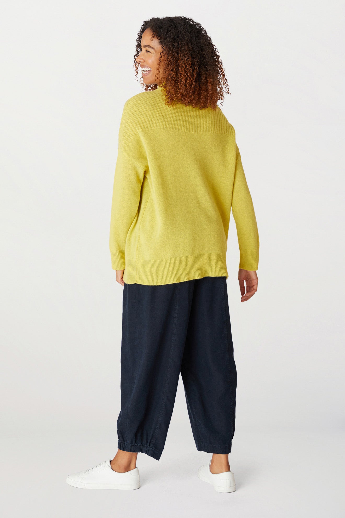 A person with curly hair wearing a bright yellow Cashmere Blend Rib Turtle Neck, dark loose-fitting pants, and white sneakers stands facing away and looking back over their shoulder with a smile.