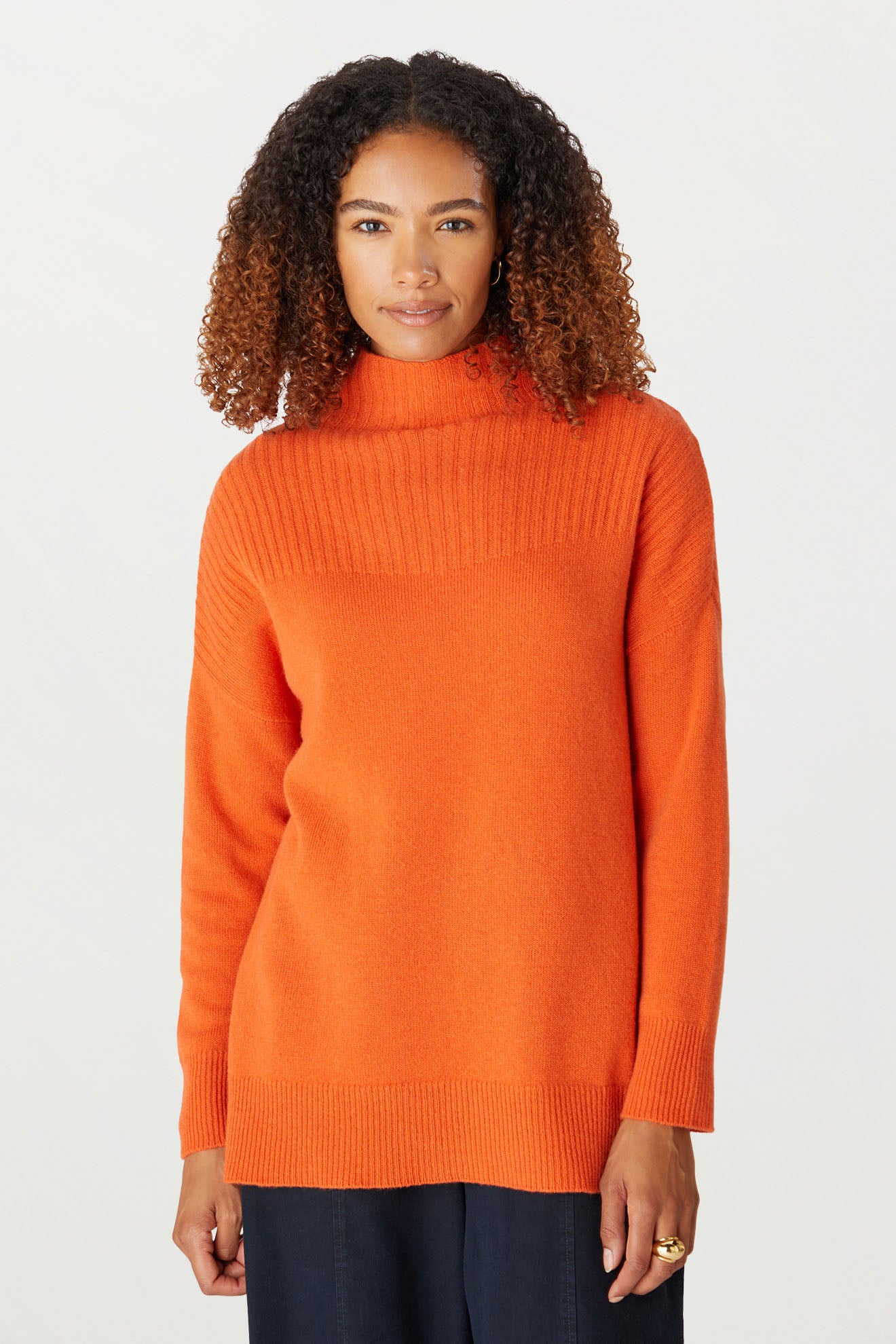 Person with curly hair wearing a Cashmere Blend Rib Turtle Neck in orange paired with dark pants, standing against a plain background.
