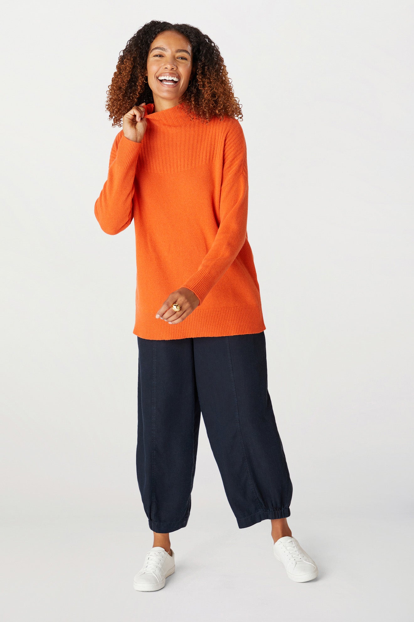 A person with curly hair is smiling, wearing a bright orange Cashmere Blend Rib Turtle Neck sweater, navy pants, and white sneakers. They are posing against a plain white background.