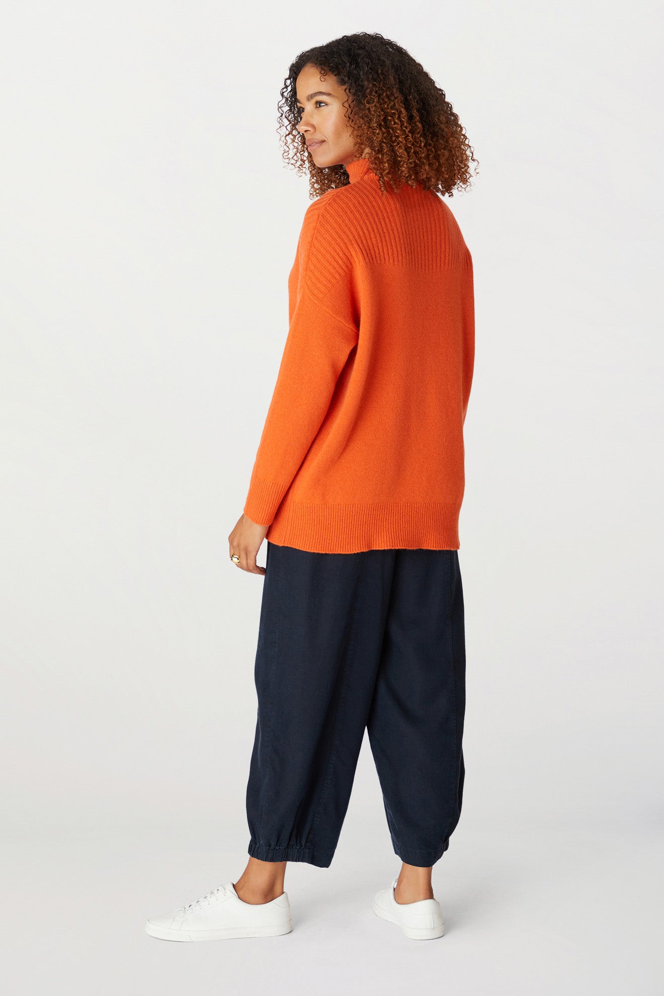 Person standing, facing away, wearing a bright orange Cashmere Blend Rib Turtle Neck—a cold-weather staple—black loose-fitting pants, and white sneakers against a plain background.