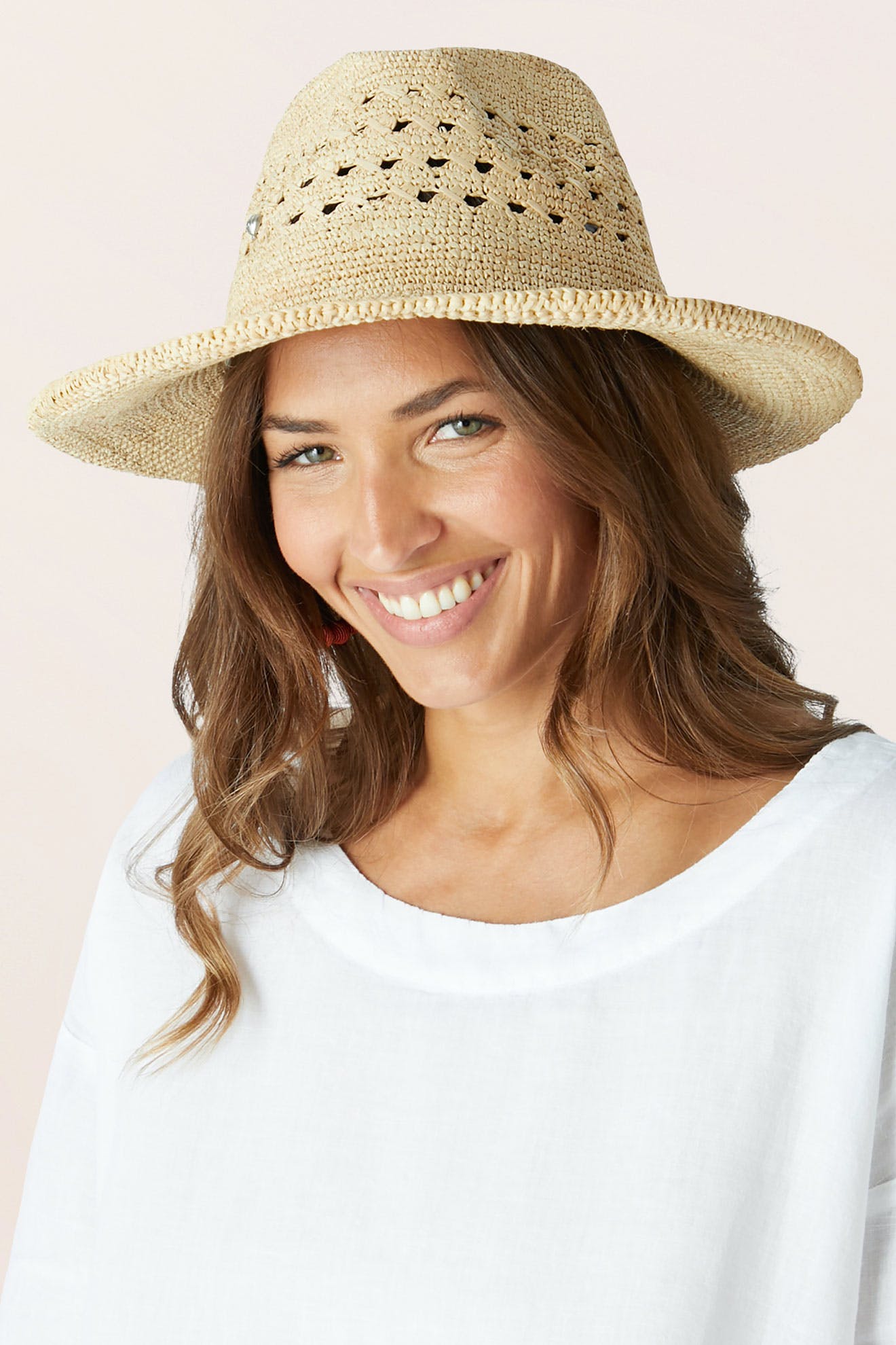A woman with long brown hair smiles at the camera, wearing the lightweight Chapeau Westosia summer hat and a loose white top against a light background.