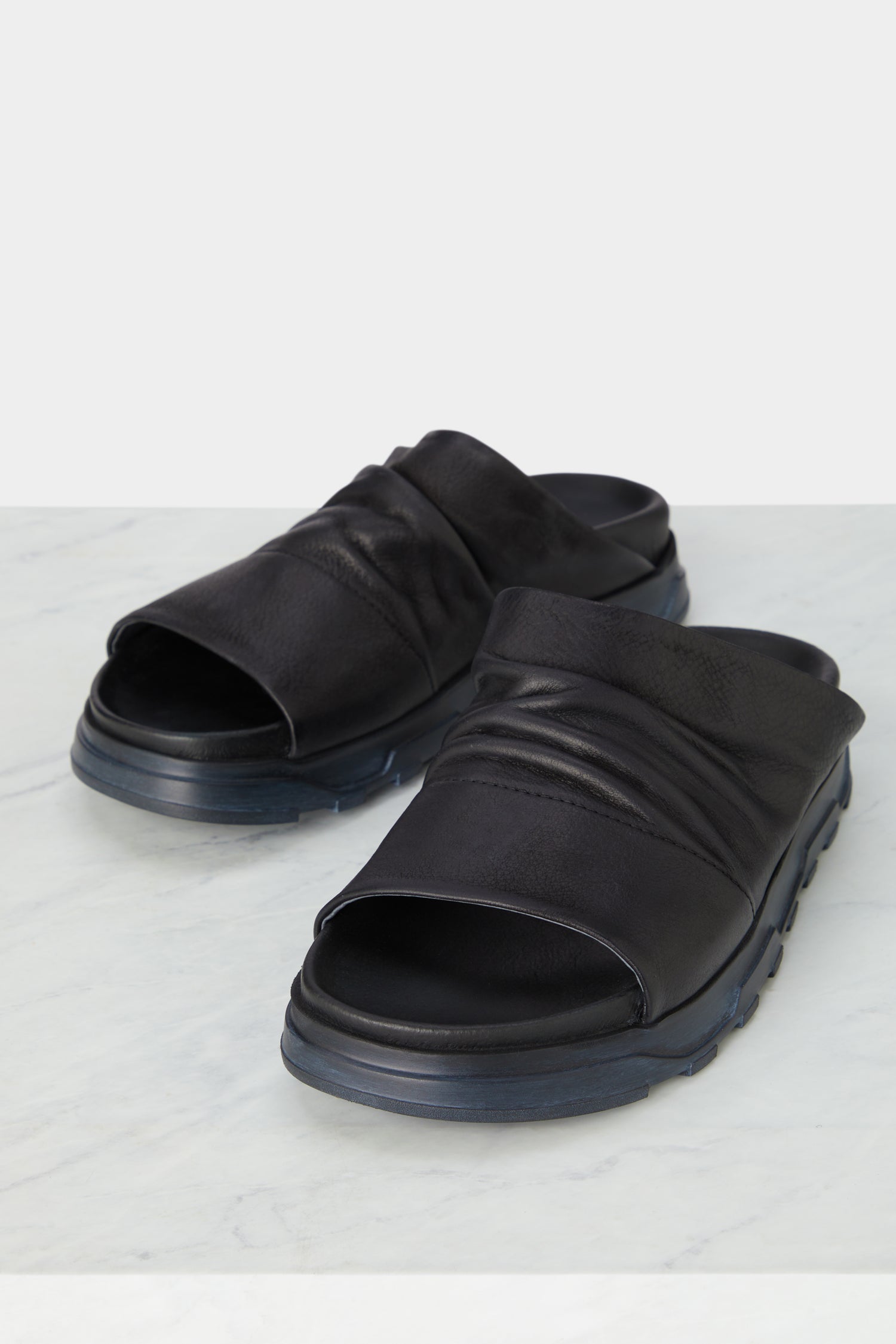 A pair of minimalist black leather Crushed Leather Sliders on a marble floor, showcasing their handcrafted quality.