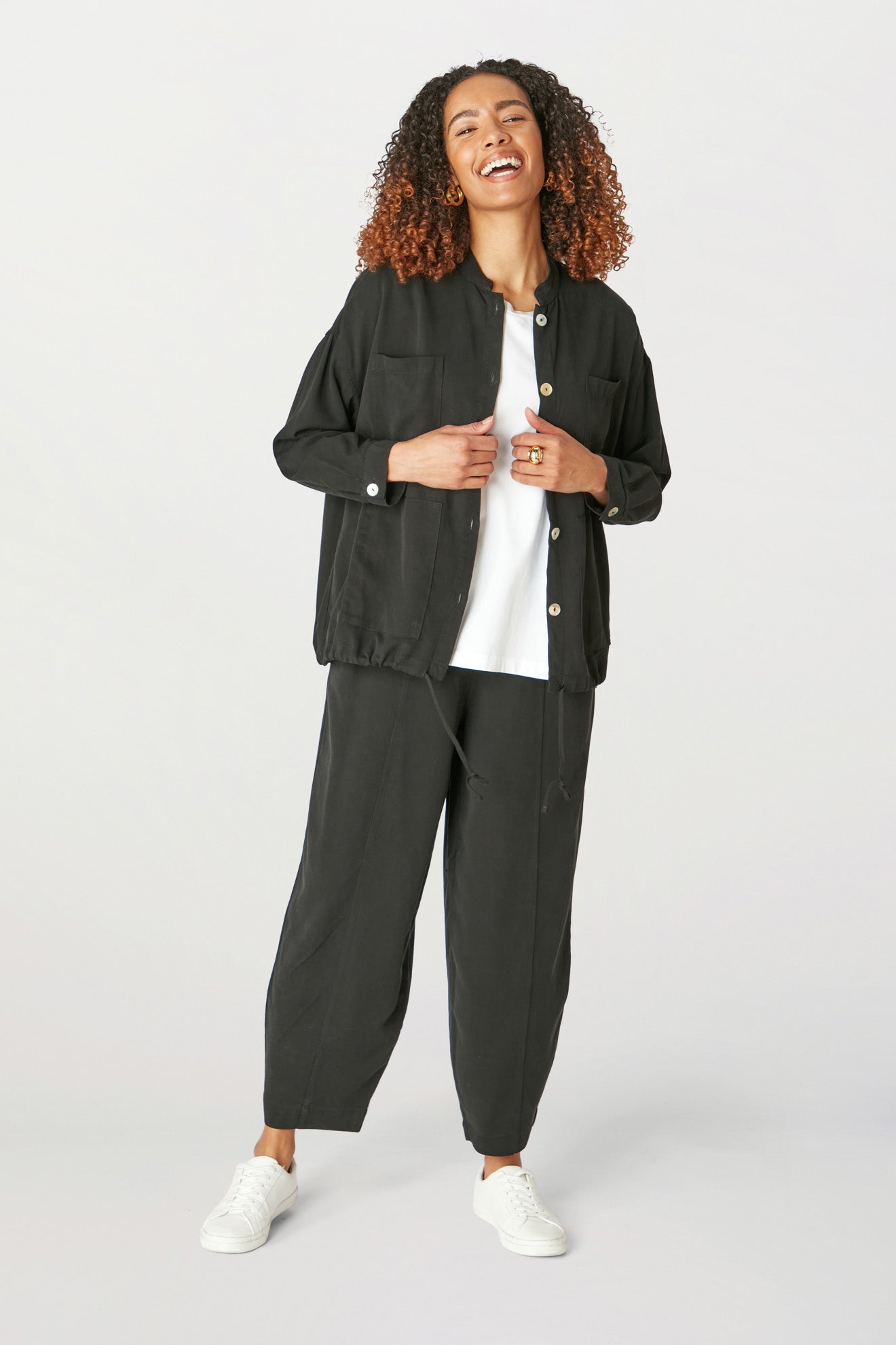 A woman with curly hair smiles, standing and wearing a Fluid Twill Drawstring Jacket, white shirt, and black trousers, completed with white sneakers.