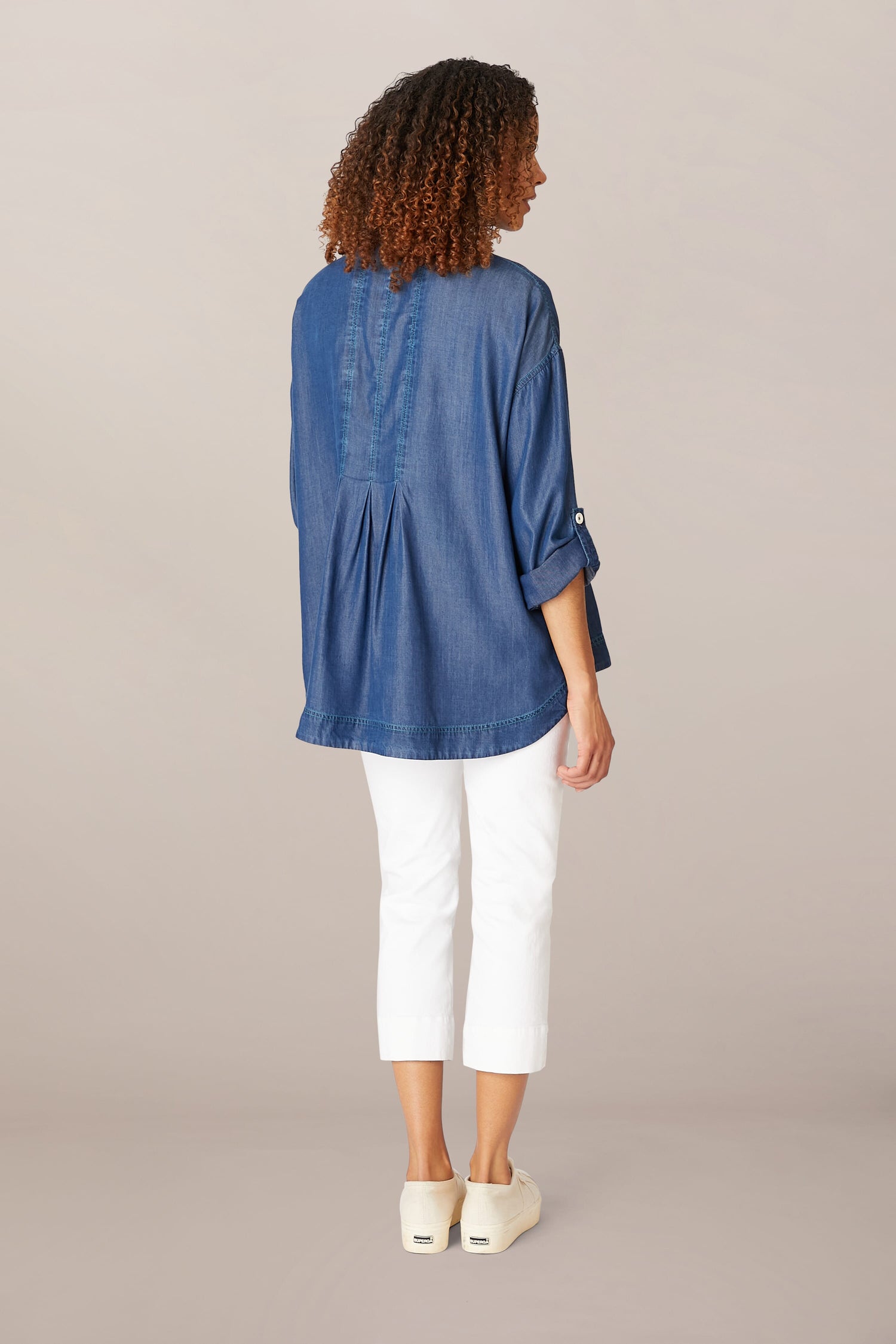Woman standing with her back to the camera, wearing a Fluid Denim Relaxed Shirt, white pants, and white shoes against a neutral background.