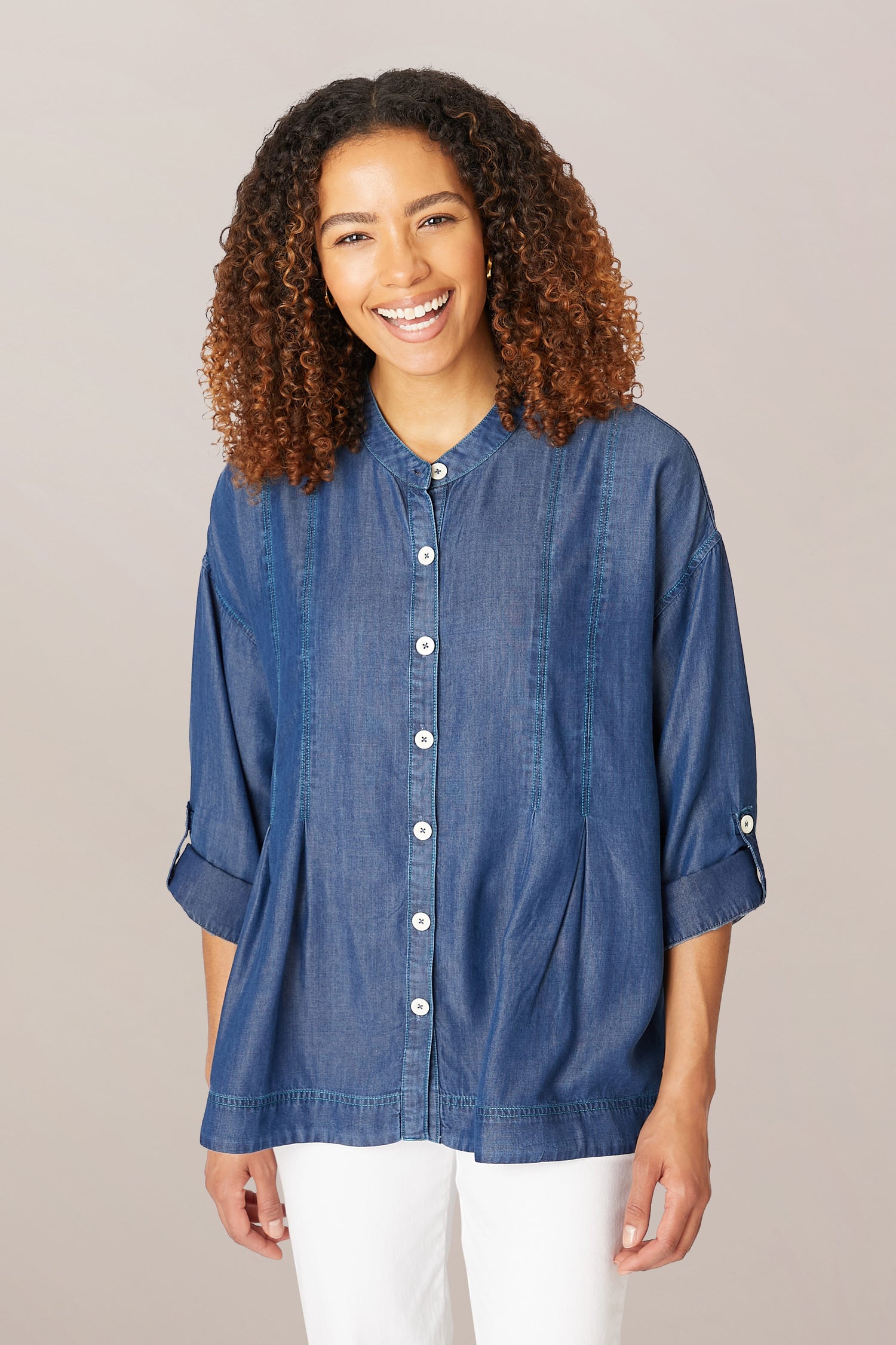 A woman with curly hair smiling, wearing a Fluid Denim Relaxed Shirt with a front button placket and white pants against a neutral background.