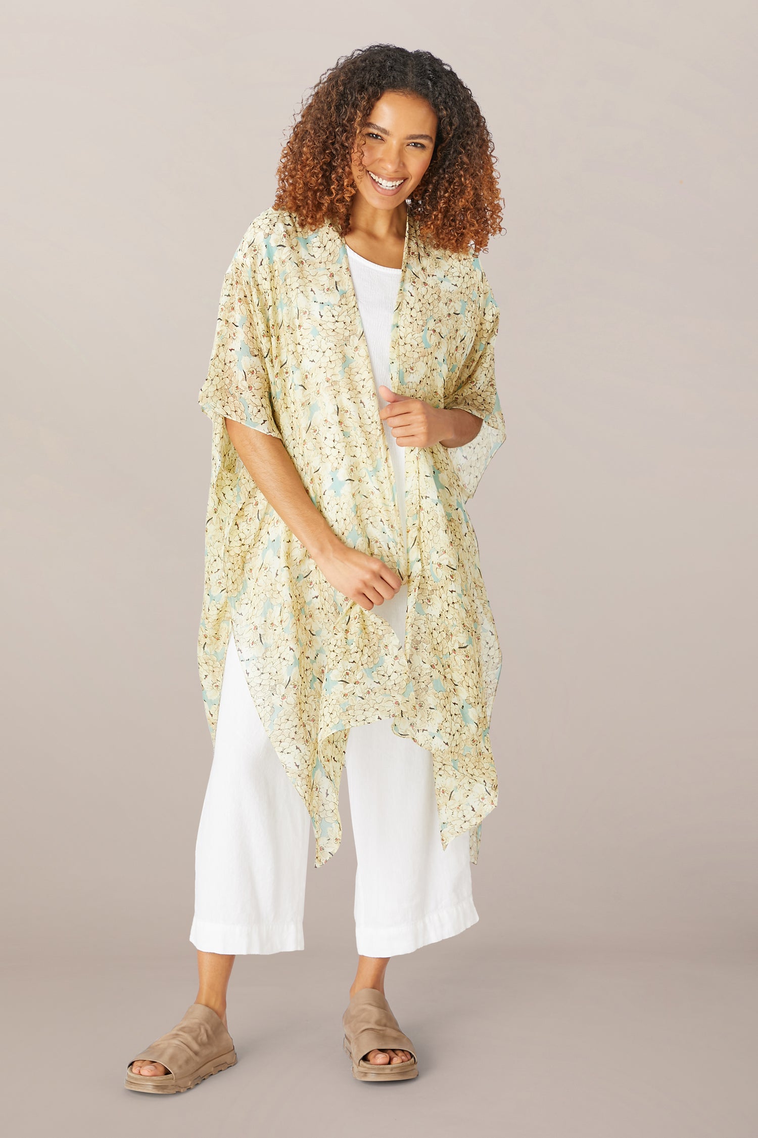 A woman smiles, wearing the Blossom Blue Throwover by One Hundred Stars over a white top and pants with beige sandals, posed against a neutral background.