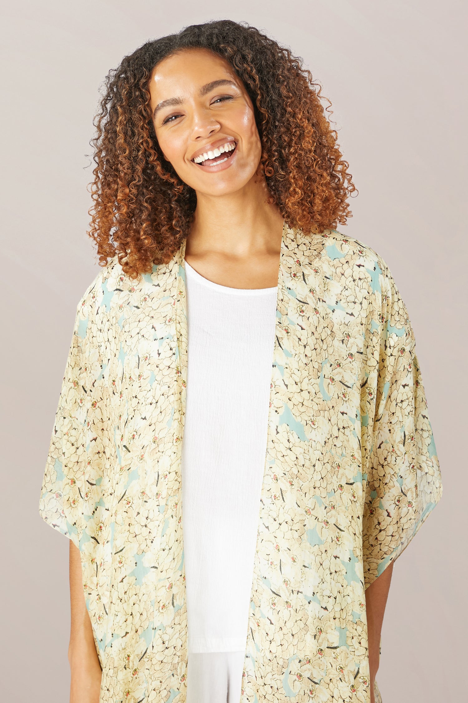 A woman with curly hair smiles, wearing the Blossom Blue Throwover—a floral-patterned kimono from One Hundred Stars—over a white top, set against a plain light background.