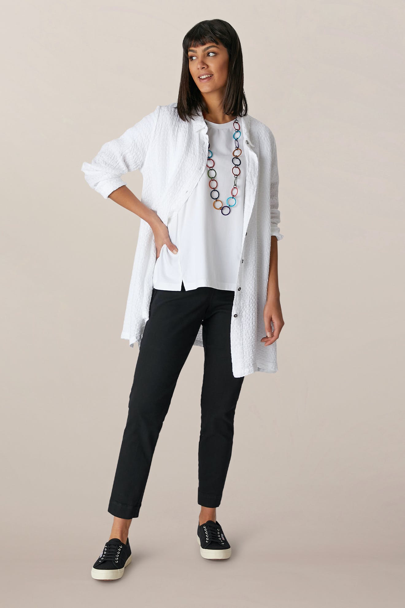 A woman stands against a plain background wearing a long white textured shirt over a white top, SS22 Stretch Cotton Trouser with an elasticated waist, black sneakers, and a colorful beaded necklace.