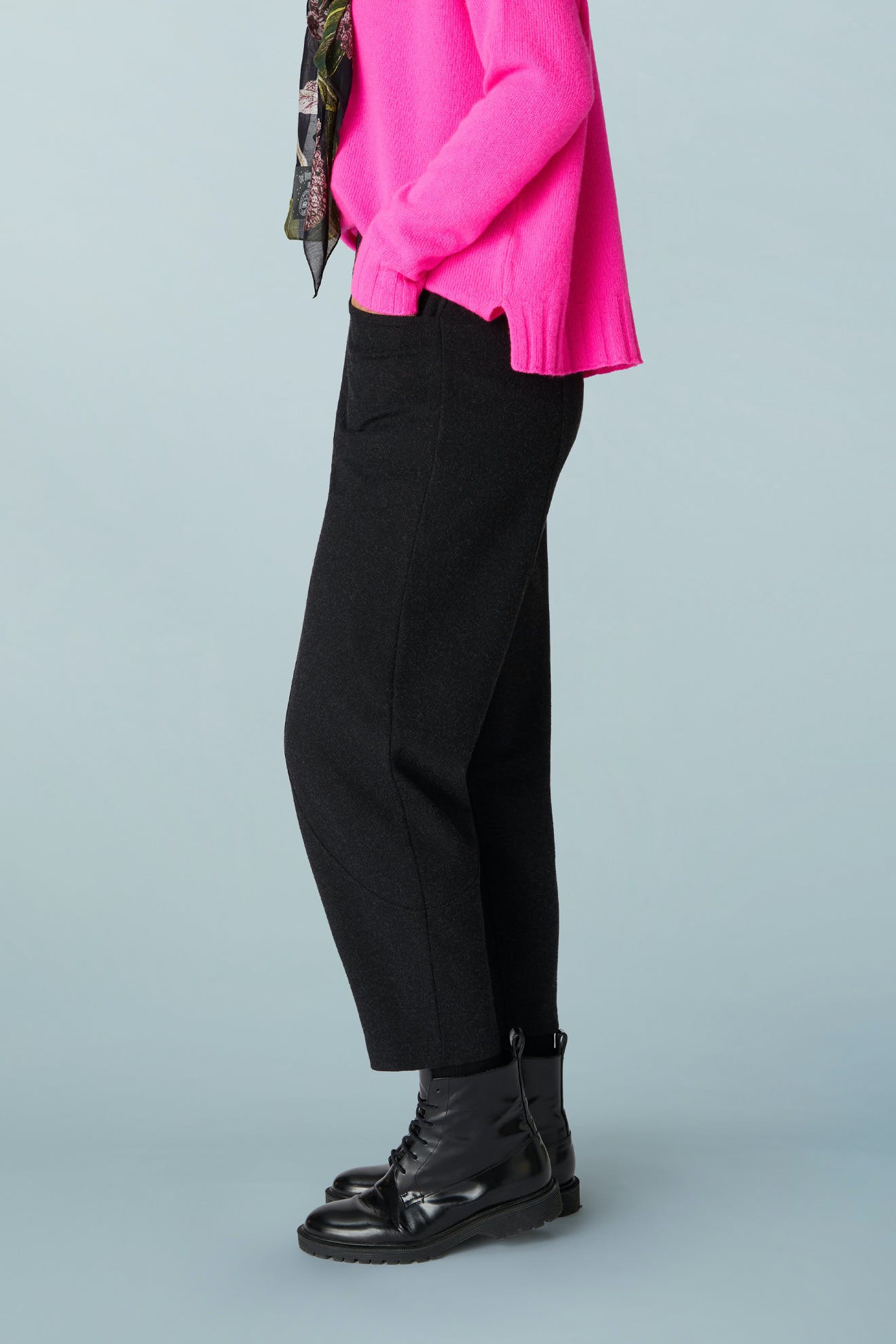 Side view of a person wearing black AW21 Marl Jersey Bubble Trouser cropped trousers with a tapered bubble silhouette, a bright pink sweater, and black lace-up boots.