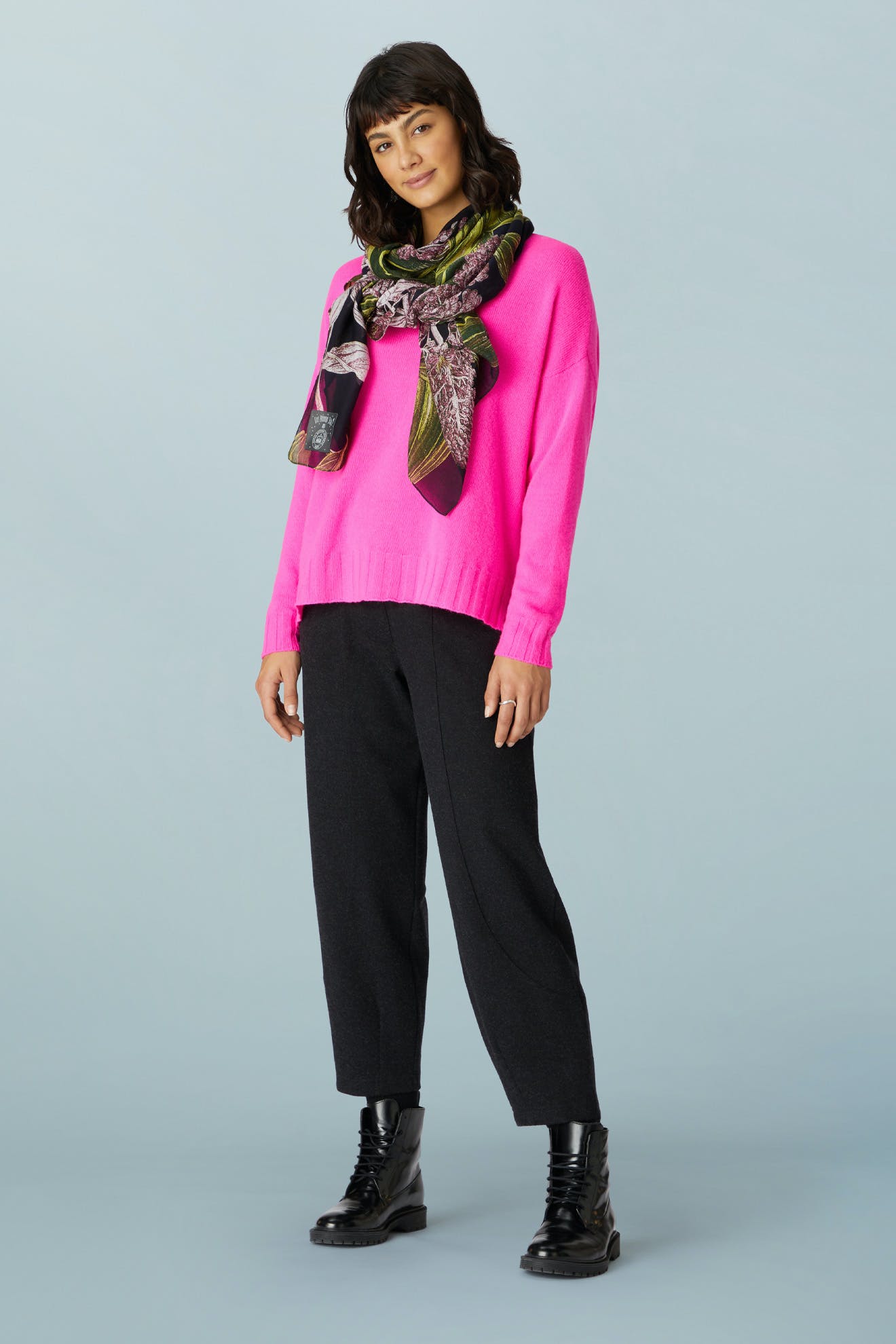 A person wearing a bright pink sweater, a patterned scarf, AW21 Marl Jersey Bubble Trouser in dark gray, and black lace-up boots.