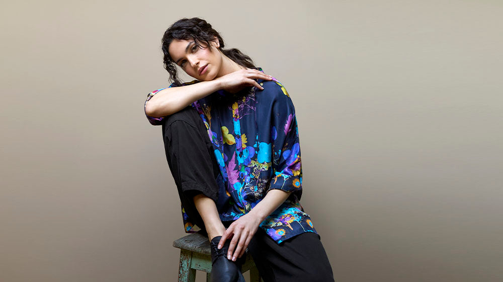 A person with wavy hair, wearing a colorful shirt and black pants, sits on a stool with one knee bent and arm resting on it against a plain background.