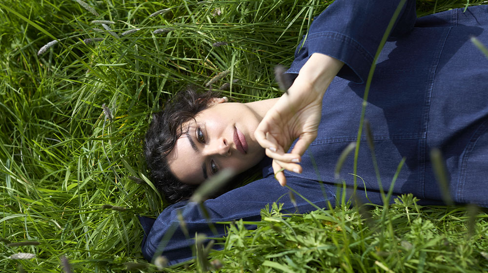 Person lying in tall grass, wearing a blue long-sleeve shirt, with one arm under their head and the other hand raised.