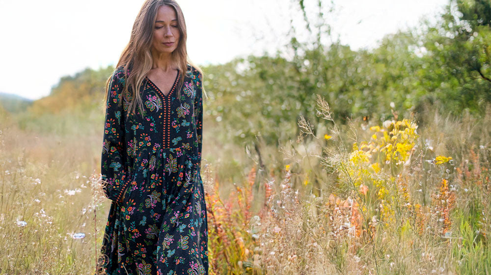 Woman in a floral dress walking through a meadow with blooming wildflowers and greenery under a clear sky.