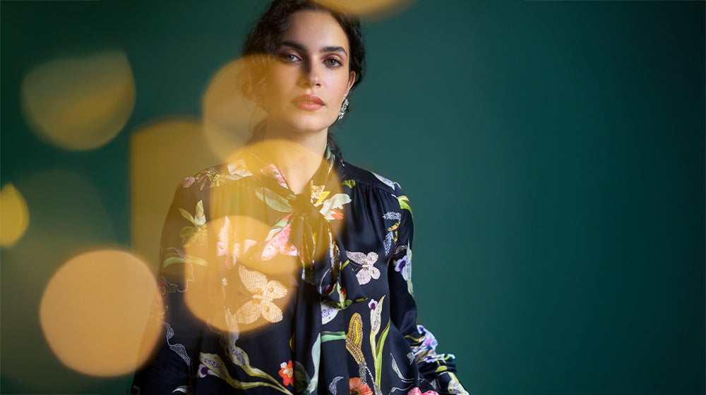 A person wearing a floral-patterned blouse stands against a green background with glowing circular lights in the foreground.