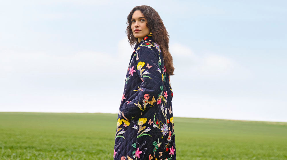 A person in a floral-patterned coat stands in a green field under a cloudy sky.