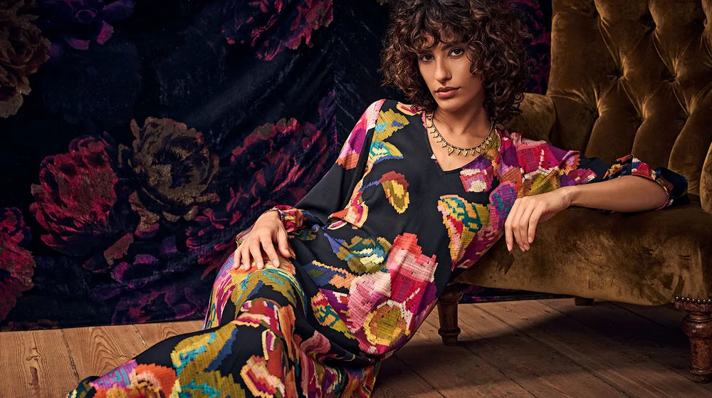 A person with curly hair wearing a long-sleeved, black dress with a colorful, abstract floral print and a gold necklace, reclining in an armchair.
