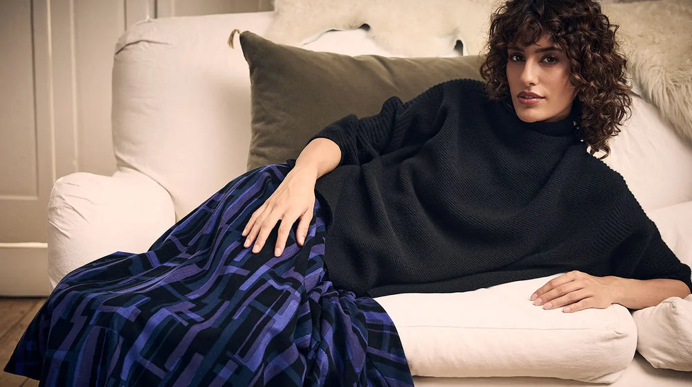 A woman with curly hair wearing a black sweater and a blue and black patterned skirt reclining on a white sofa.