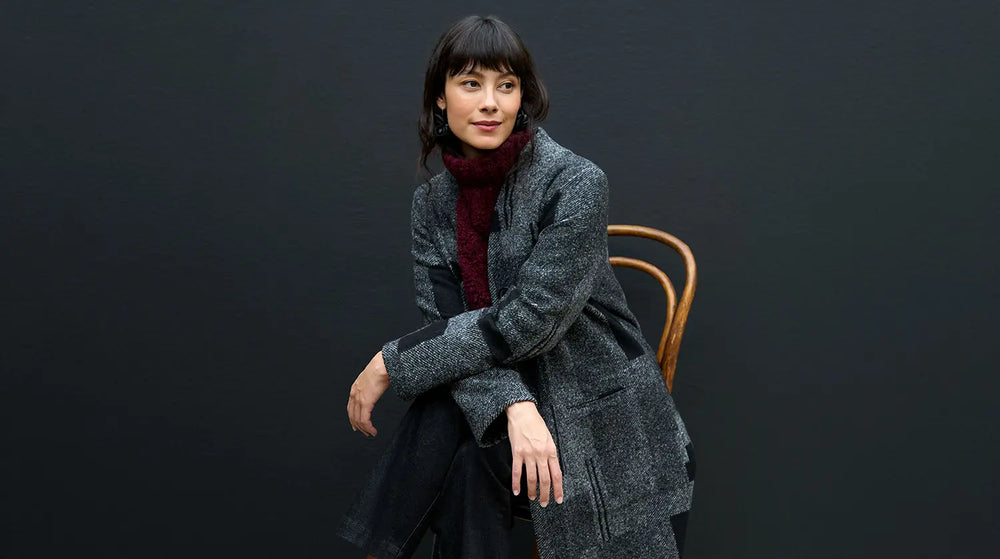 A woman wearing a dark grey coat and a burgundy scarf sits on a wooden chair against a dark background.