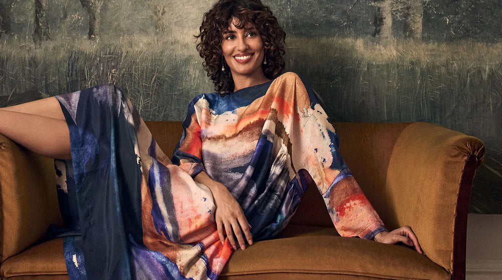 A smiling woman with curly hair wearing a long-sleeved dress with a multi-colored abstract landscape print, reclining on a brown sofa.