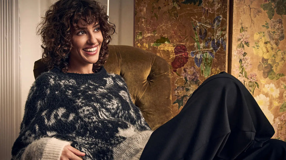 A smiling woman wearing a dark patterned sweater sits in an armchair.
