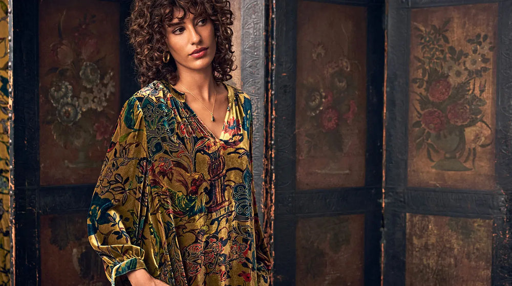 A woman with curly hair wearing a long-sleeved, gold-toned velvet dress with a multicolor floral and paisley pattern.