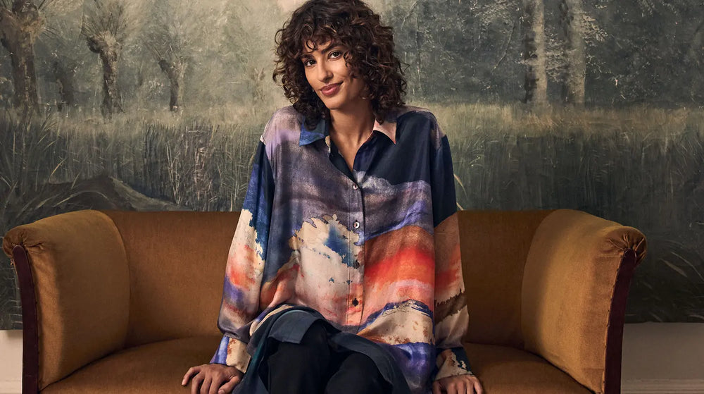 A person with curly hair sits on a brown sofa wearing a button-down shirt featuring an abstract landscape print of a sunset over water.