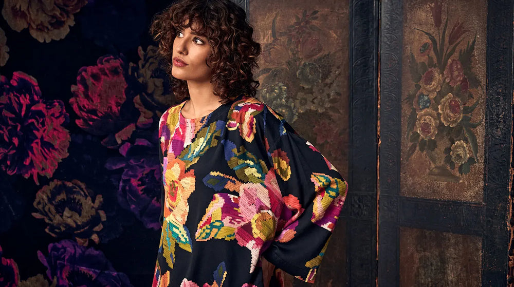 A woman with curly hair wears a long-sleeved dress with a dark background and a colorful, pixelated floral pattern.