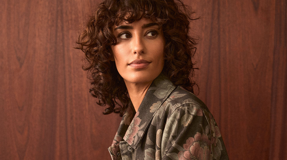 A person with curly brown hair wearing a floral-patterned jacket looks to the side against a wooden background.