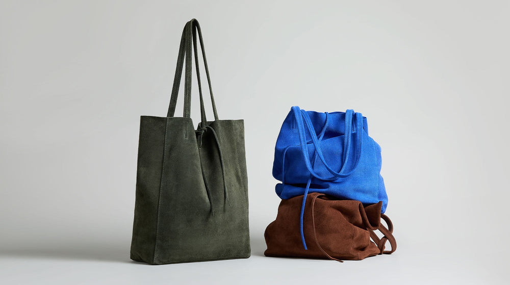 A dark green suede tote bag stands upright next to two slouchy suede drawstring bags, one blue stacked on top of one brown, against a plain light background.
