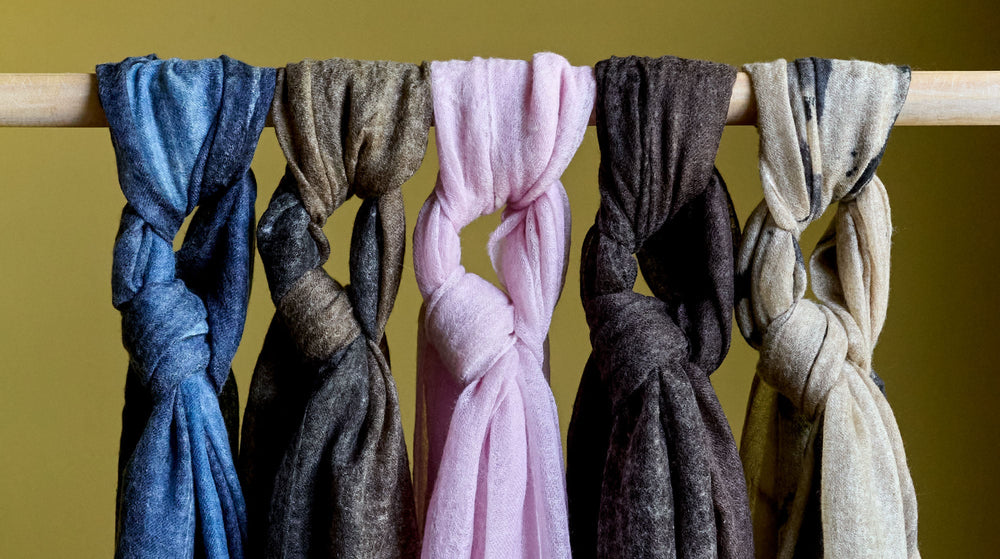 Five scarves in blue, brown, pink, dark gray, and beige colors are knotted over a wooden rod against a plain yellow background.