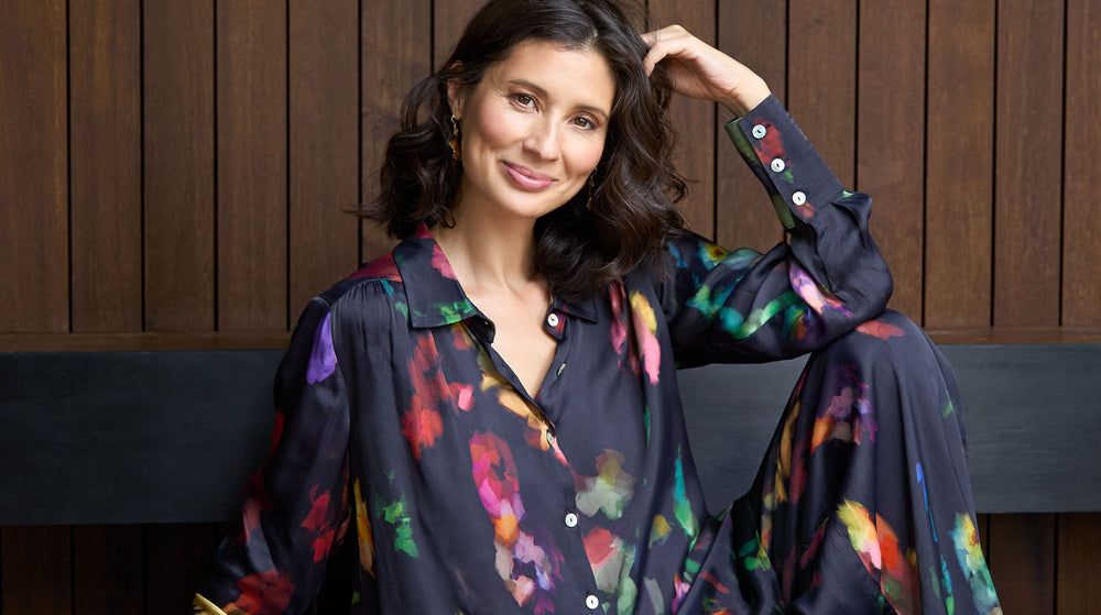 A woman with wavy dark hair sits in front of a wooden wall, smiling, wearing a loose-fitting, dark floral-patterned shirt and pants.
