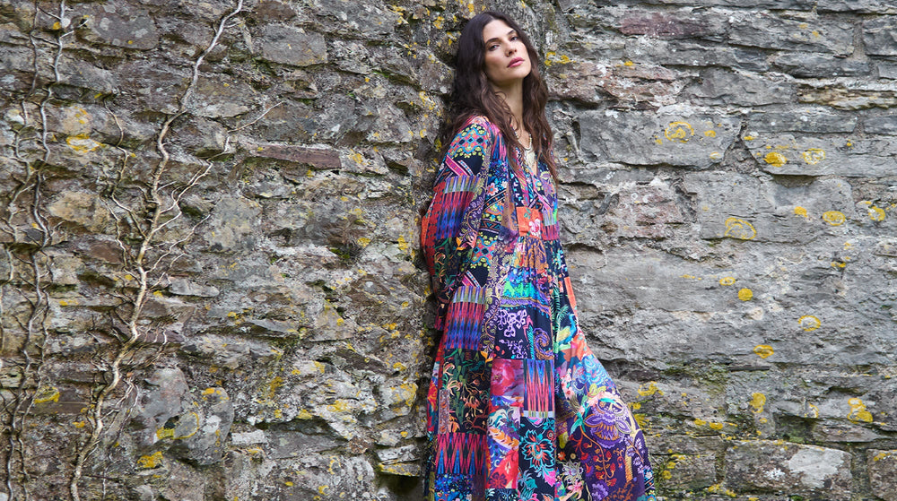 A woman wearing a long, colorful patterned dress stands against an old stone wall with patches of moss and yellow lichen.