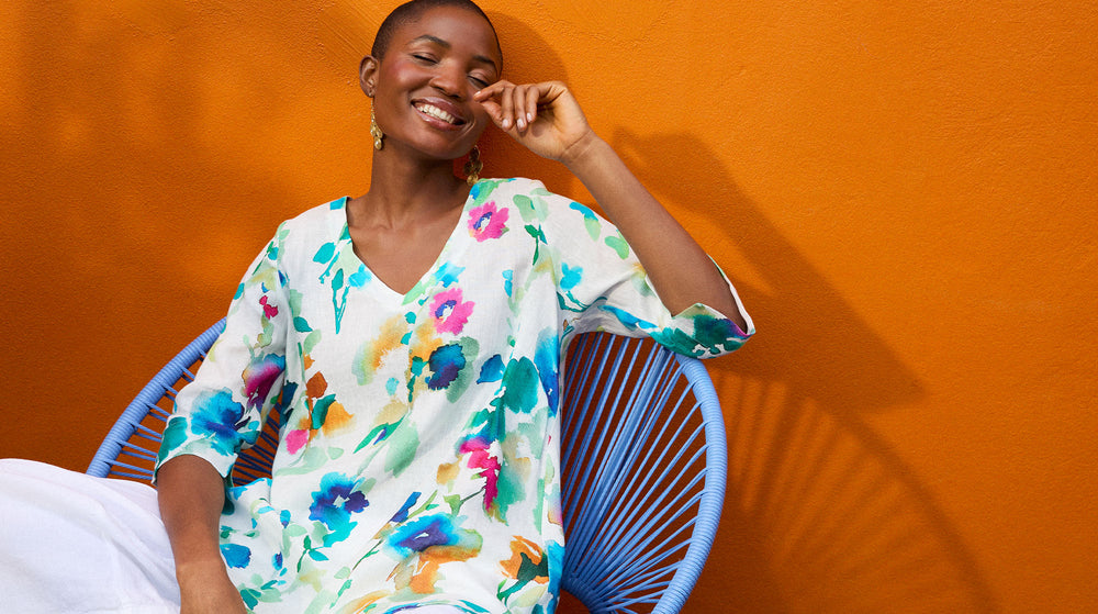 Person in a floral blouse sits on a blue chair against an orange wall, smiling.