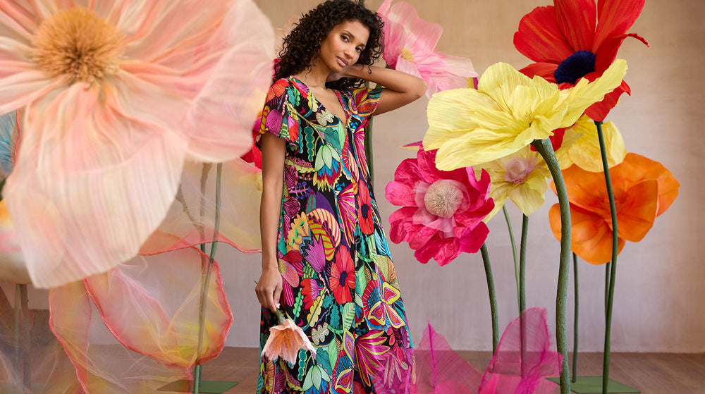 A woman wearing a colorful, patterned dress stands among large, artificial flowers in various bright colors against a neutral background.