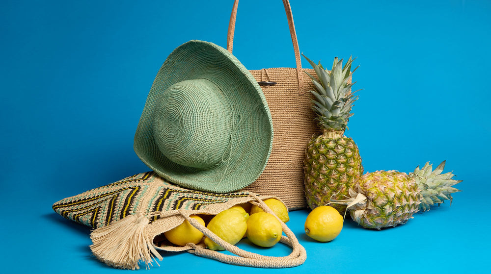 A straw tote bag, green sun hat, patterned clutch, pineapples, and several lemons are arranged together against a bright blue background.