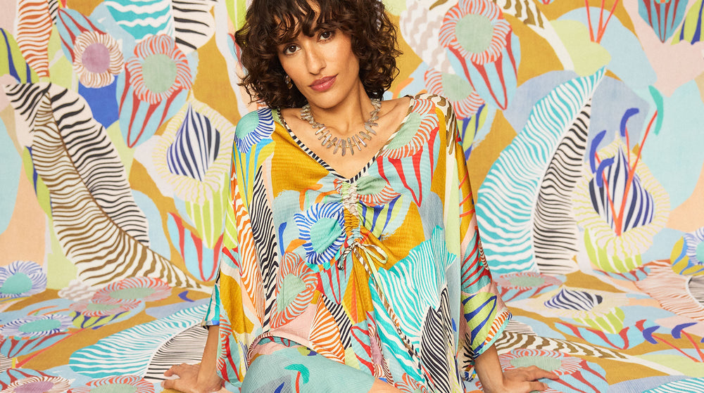 A woman in a colorful, patterned outfit sits against a matching abstract, multicolored background with stripes and floral shapes.