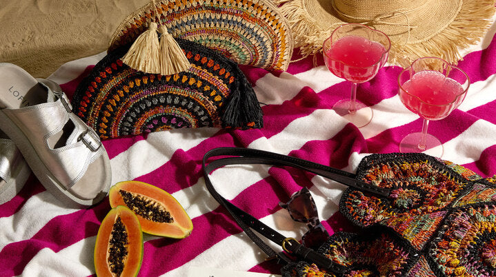 Straw hat, silver sandal, crochet handbags, two pink drinks in glasses, papaya slices, and a striped towel arranged on a surface.