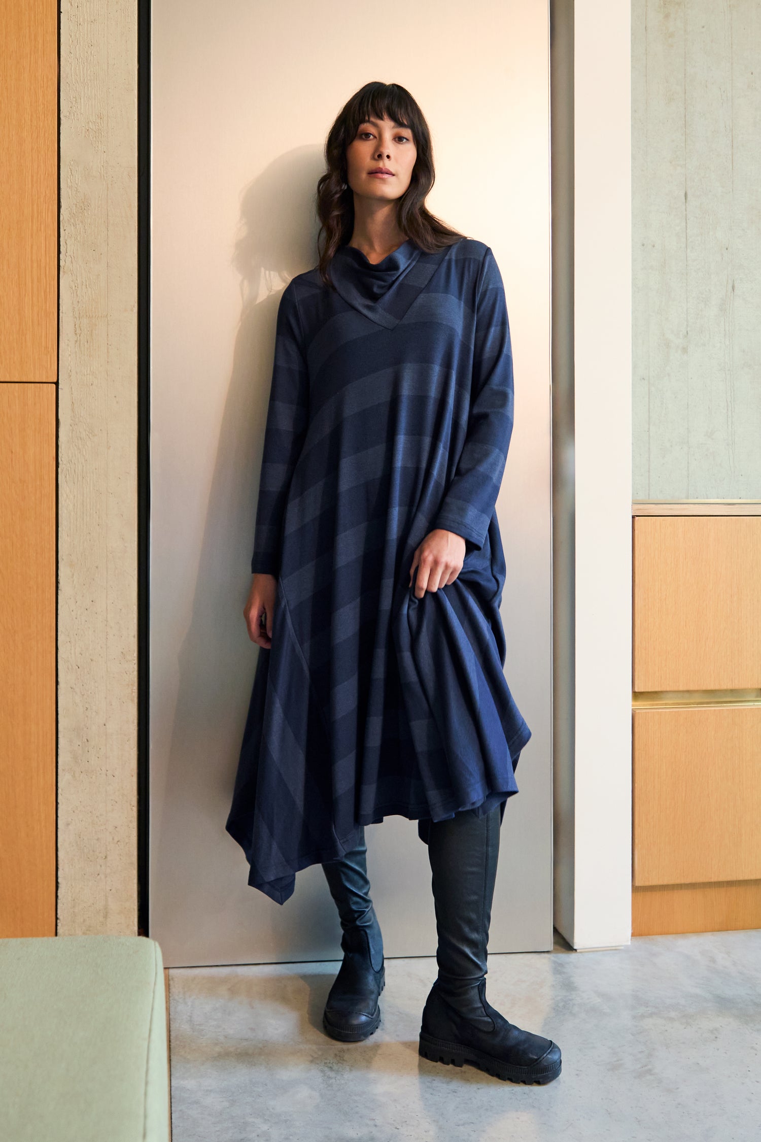 Indoors, a person wears the Soft Viscose Jersey Stripe Dress—a loose-fitting, dark blue patterned jersey dress with an asymmetric hem—styled with black boots and holding part of the dress.