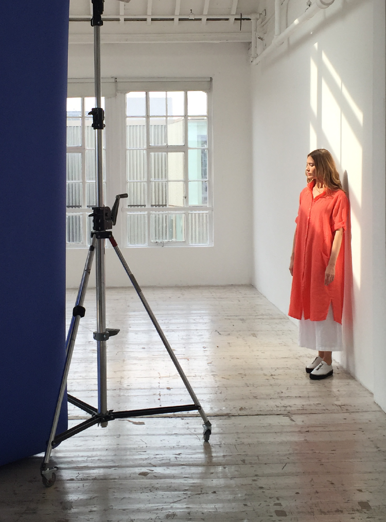 woman wearing a long red shirt and white trousers stood in a photography studio
