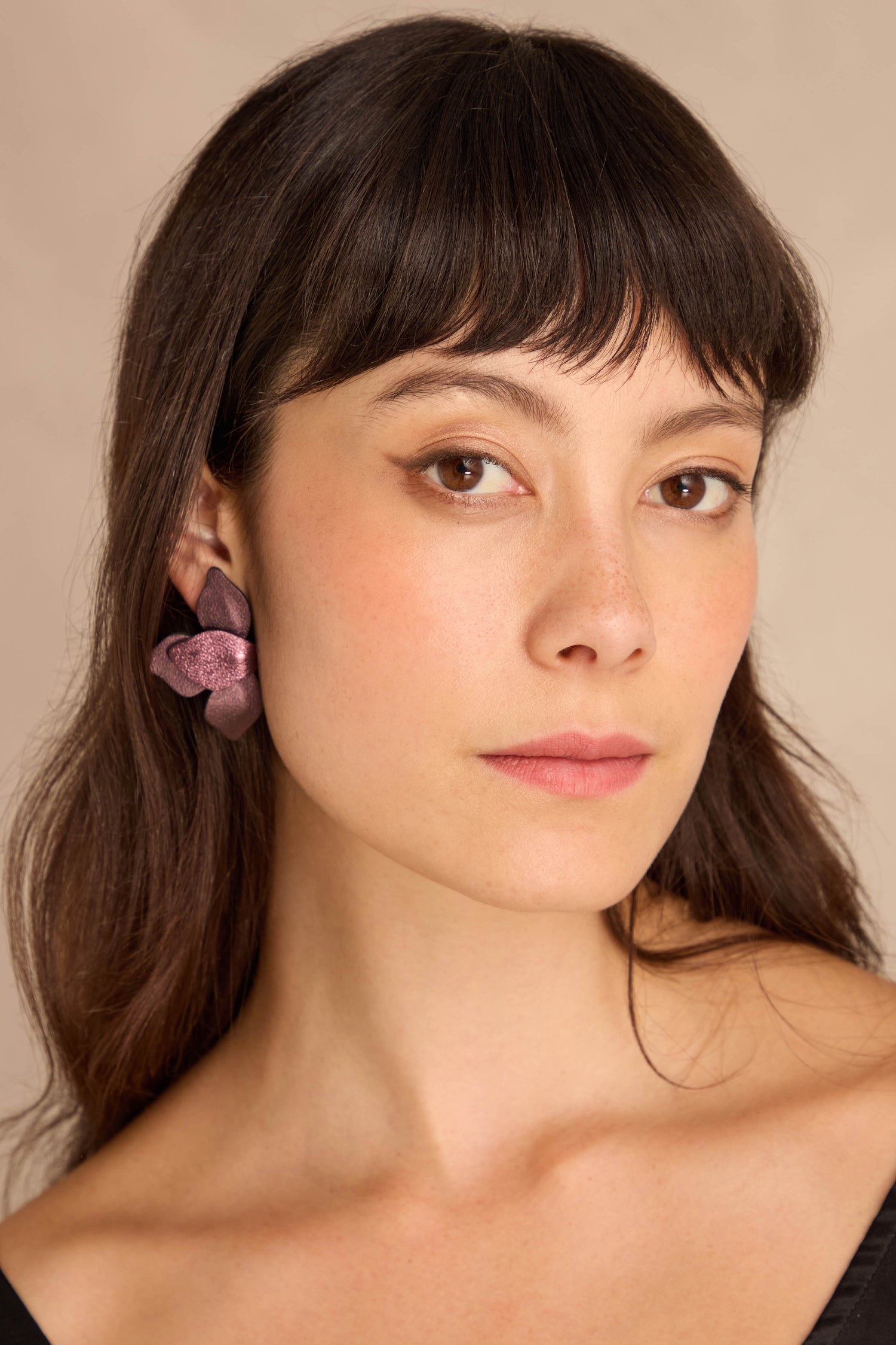 Woman with straight brown hair and bangs wears a black top and Metallic Burgundy Leather Petal Earrings on her left ear, looking at the camera.