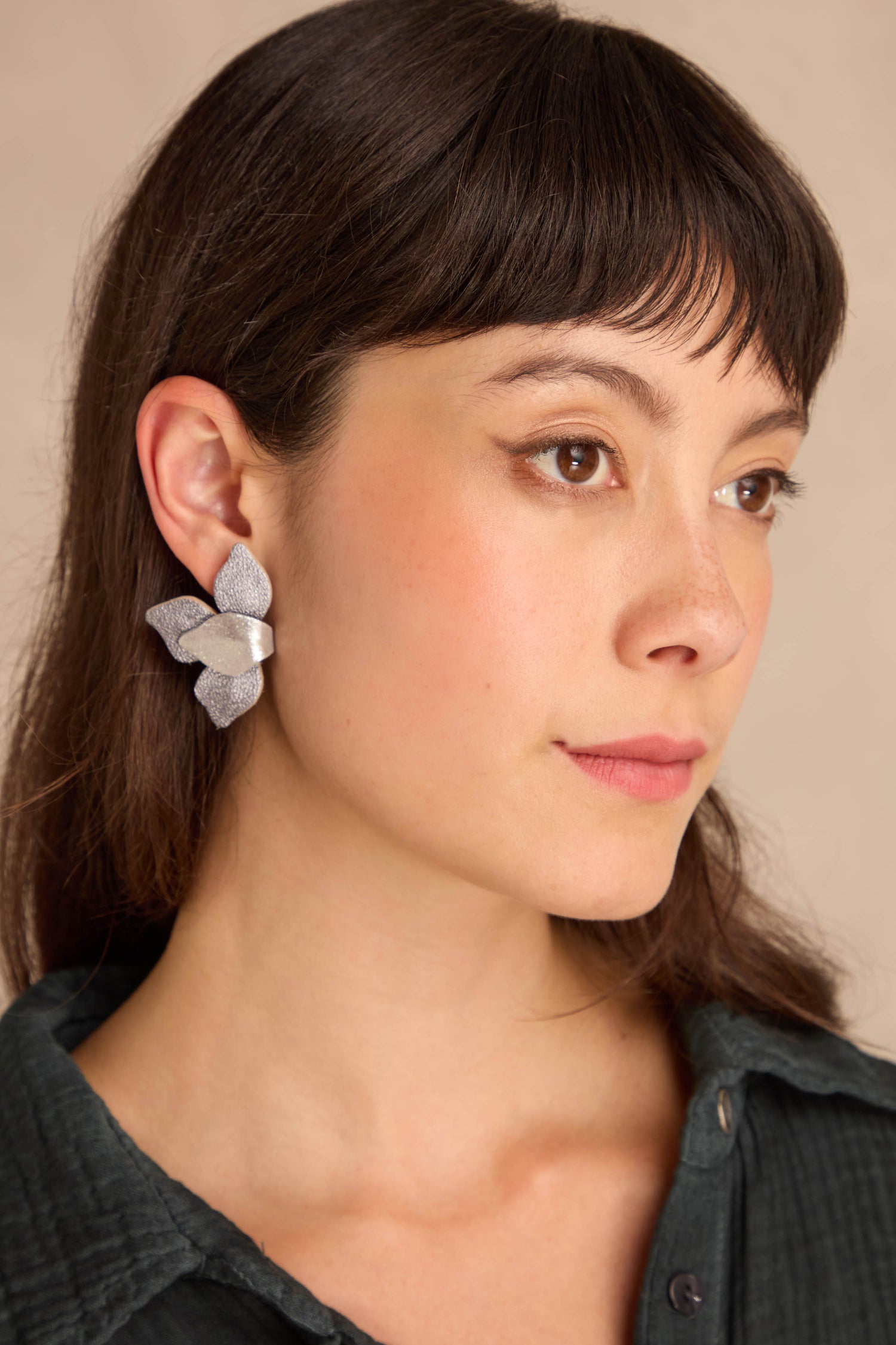 Woman with straight brown hair in a dark green shirt wears Metallic Grey Leather Petal Earrings, gazing to the side against a neutral background.