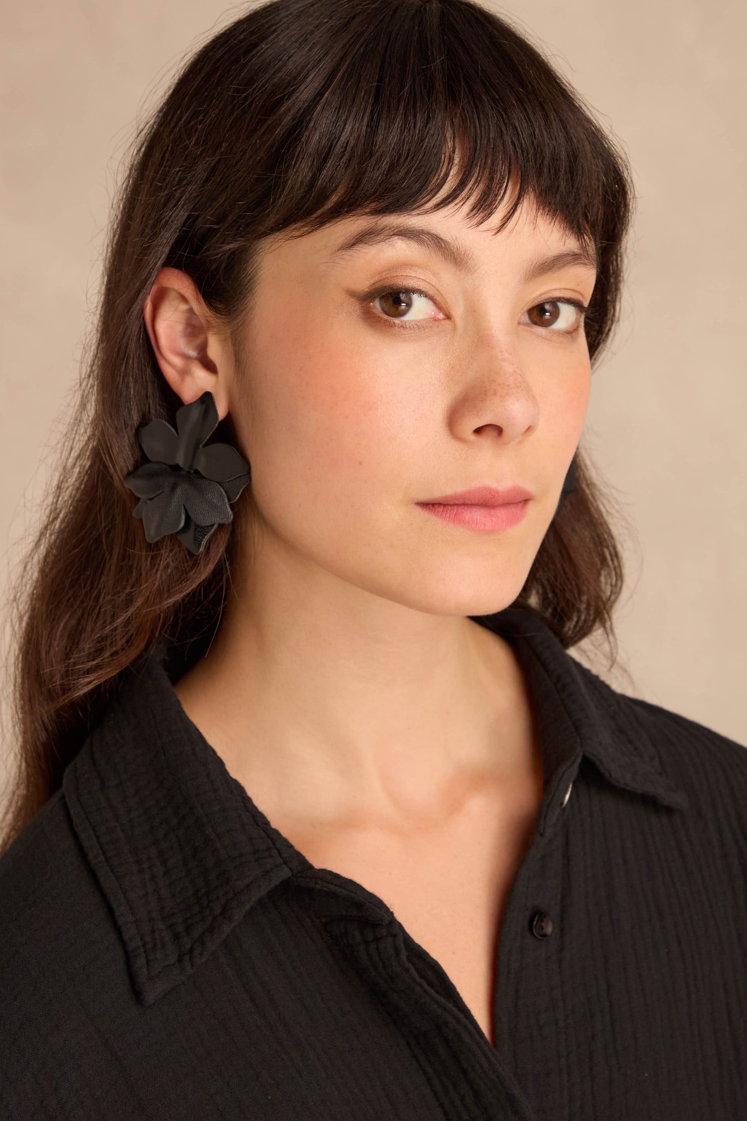 Woman with long brown hair and a black collared shirt wears Metallic Leather Flower Earrings, looking at the camera against a neutral background.