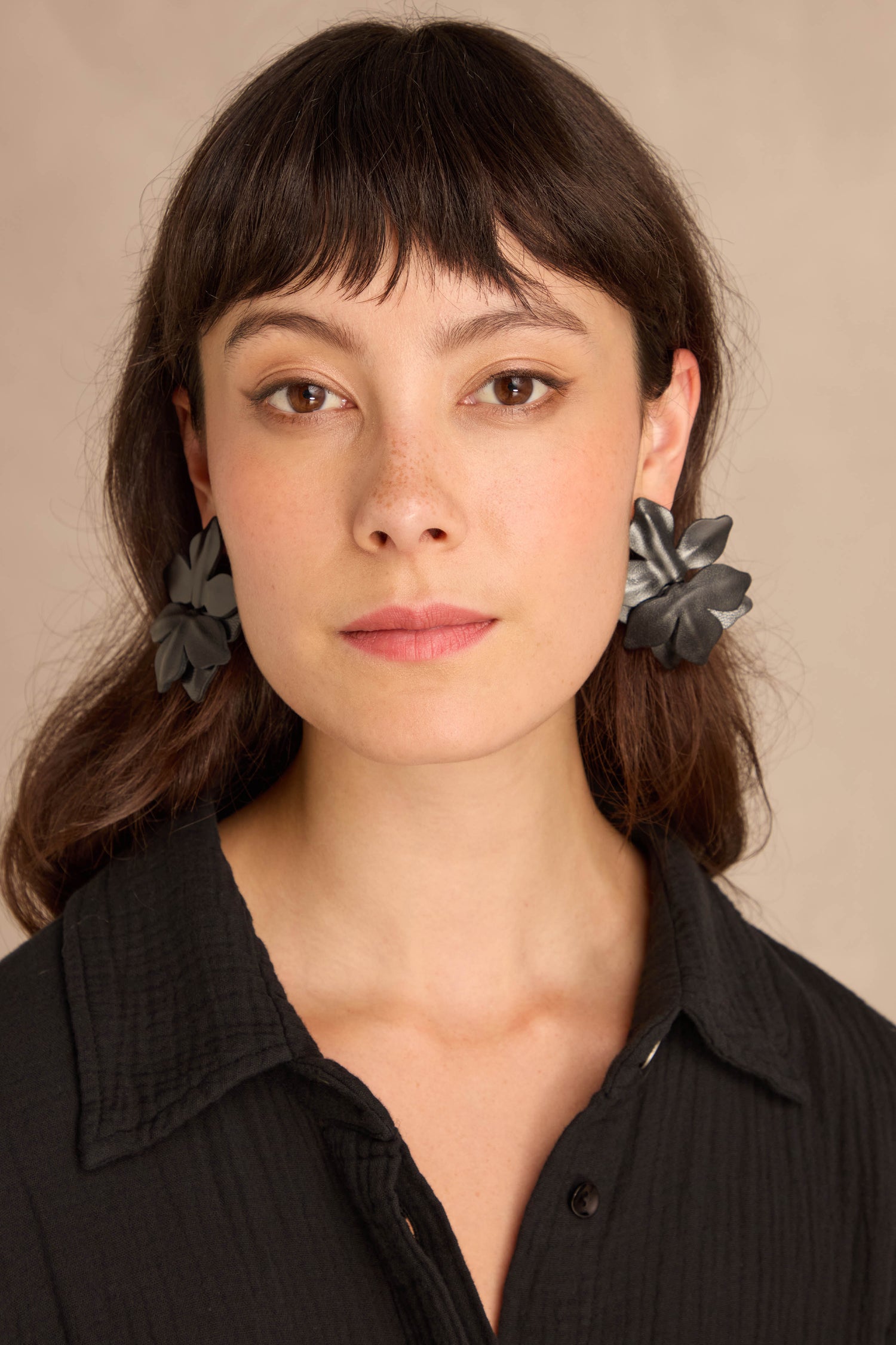 Woman with straight brown hair wears a black textured blouse and Metallic Leather Flower Earrings, posing against a neutral backdrop.