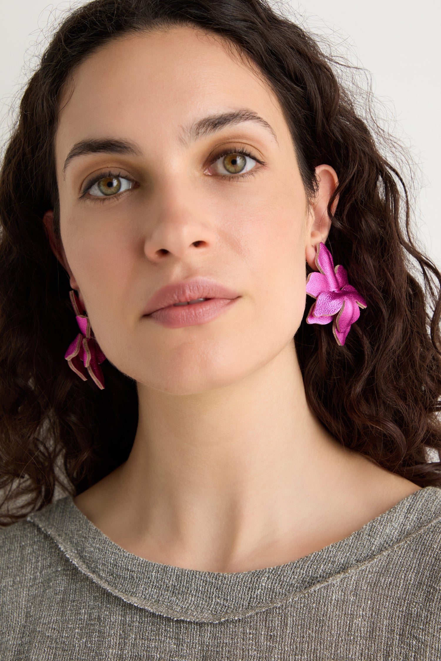Woman with curly brown hair wears bright pink Metallic Leather Flower Earrings in a flower shape and a grey top, facing forward against a plain background.