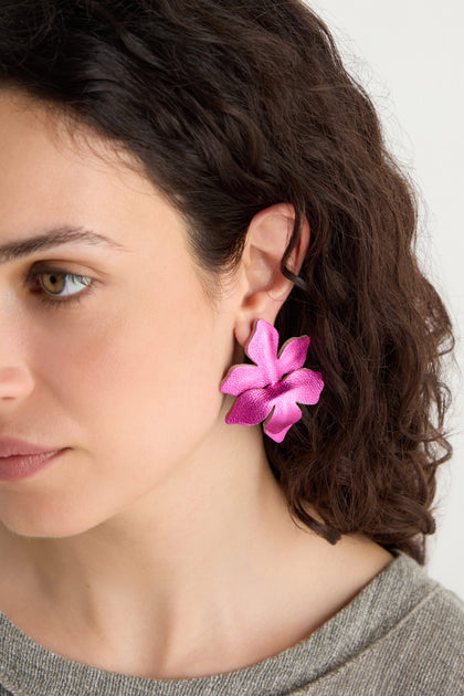 A woman with curly brown hair wears a Metallic Leather Flower Earring in metallic fuchsia, handcrafted in a large flower design, on her left ear.